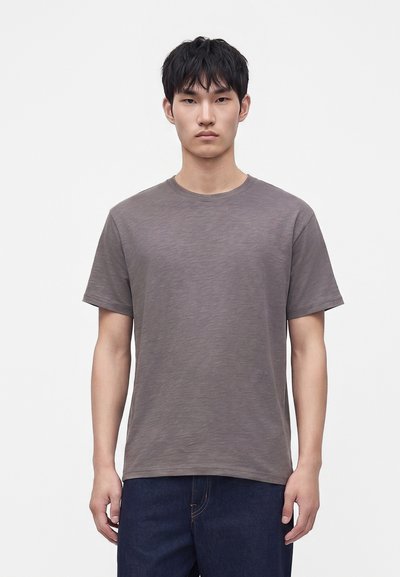 Jeune homme aux cheveux courts noirs portant un t-shirt gris uni à manches courtes et un jean bleu foncé sur fond blanc.