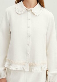 Mujer vistiendo una blusa crema con botones, cuello con volantes, borde de encaje y dobladillo con volantes, mostrada desde el cuello hasta las caderas.