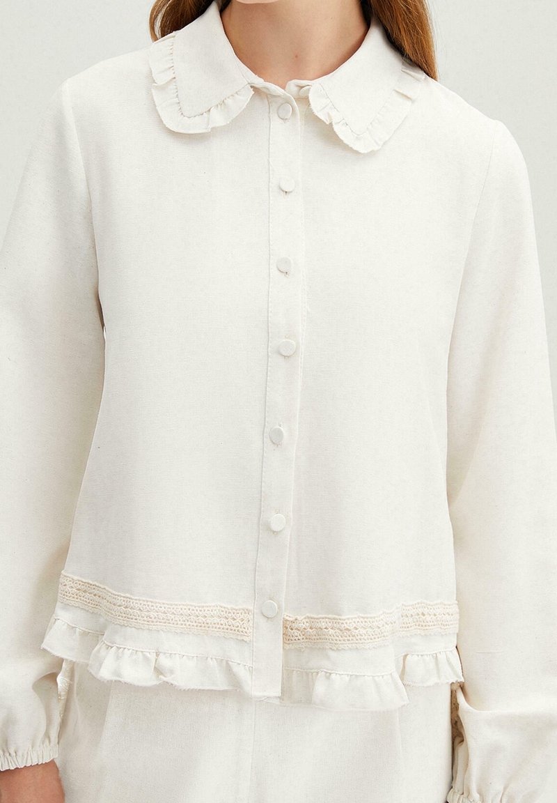 Mujer vistiendo una blusa crema con botones, cuello con volantes, borde de encaje y dobladillo con volantes, mostrada desde el cuello hasta las caderas.