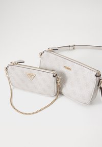 Zwei passgenaue Taschen aus hellbeigem Stoff mit einem geprägten Logo-Muster. Hardware und Reißverschlüsse in Goldoptik, eine mit abnehmbarem Kettenriemen.