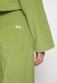 Groene corduroy-outfit met verticale ribbeltextuur, met een label op de achterzak en brede mouwen.