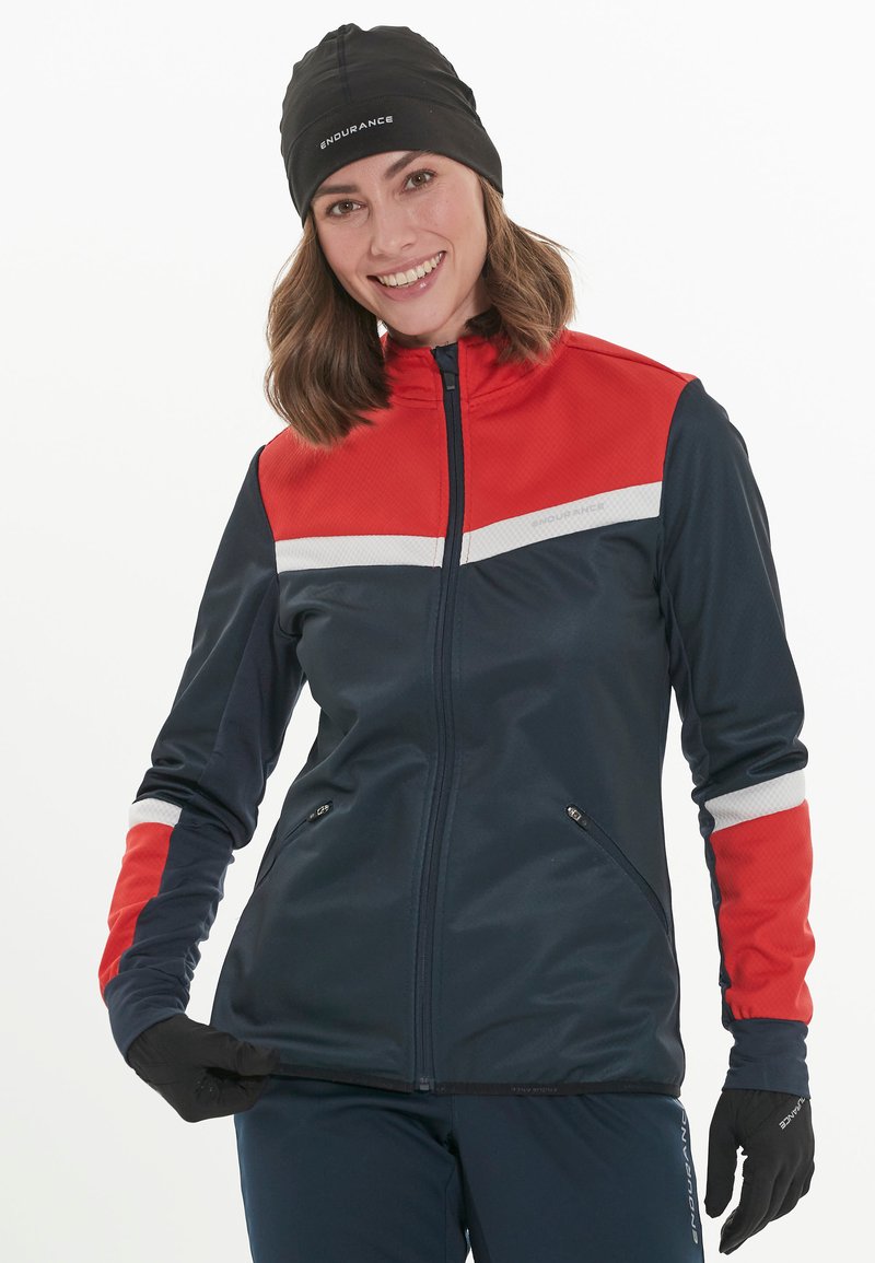 Marineblauwe en rode zip-up jas met witte accenten; gemaakt van lichtgewicht materiaal, met een hoge kraag en gecapte zijzakken. Zwarte handschoenen.
