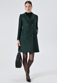 Cappotto doppiopetto verde scuro con colletto, dotato di due tasche e grandi bottoni. Abbinato a collant neri, scarpe e una borsa nera.