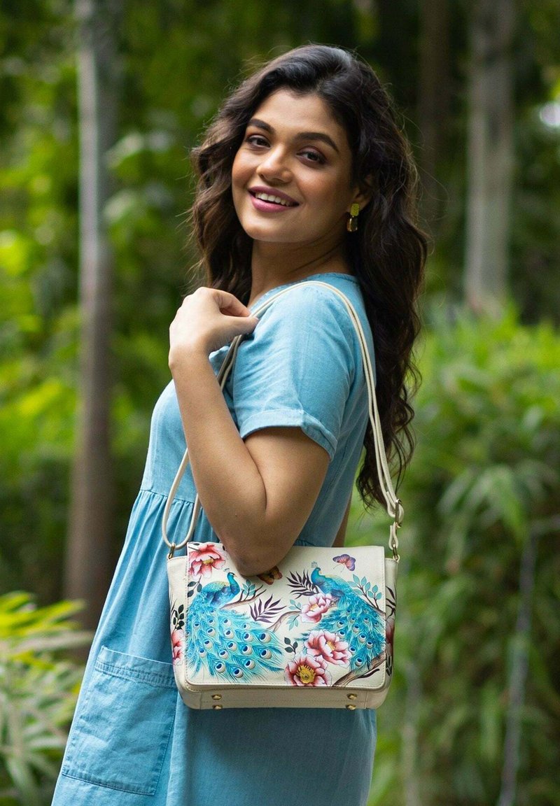 Weiße Handtasche mit Pfau- und Blumenmustern, goldfarbener Hardware und einem langen Tragegurt. Das Model trägt ein hellblaues Kleid mit kurzen Ärmeln.