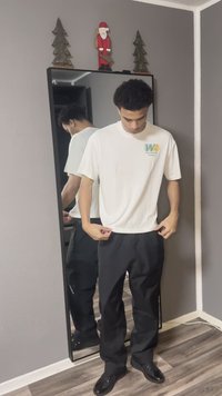 Balta T-krekls ar grafiku "Worldwide Hoops", kombinēts ar melnām, brīvi pieguļošām biksēm un melnām kurpēm, atspoguļots pilna auguma spogulī.