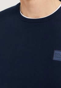 Mörkblå crewneck-tröja som bärs över en vit undertröja, med en liten rektangulär "Polo Club"-patch på bröstet.