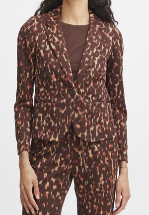 Vrouw die een bruin, aangesloten blazer en bijpassende broek draagt met een rood-beige dierenprintpatroon over een bruin topje.