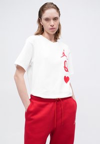 Fehér pamut póló, piros nyomott grafikákkal, beleértve egy Jumpman logót, szöveget és egy szívet. Pirossal húzott derekú melegítőnadrággal párosítva.