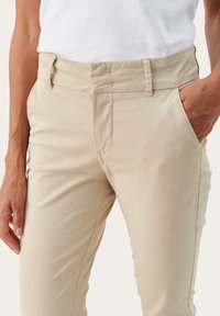 Pantaloni in cotone beige con design a gamba dritta, caratterizzati da tasche frontali e chiusura con zip, texture liscia e dettagli di cucitura discreti.