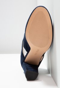 Chaussure à talon haut en daim bleu marine avec une semelle en caoutchouc texturée et des côtés ouverts, avec une semelle intérieure beige lisse et un branding discret.