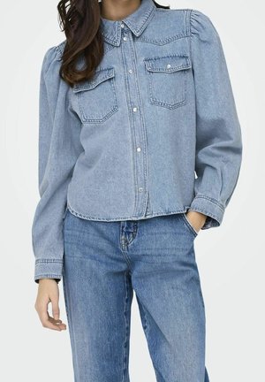 Frau trägt hellblaue Denim-Bluse mit Puffärmeln und passende blaue Denim-Jeans, Hand in der Tasche.