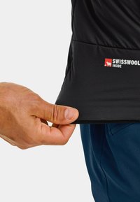 Schwarze isolierte Jacke mit einem Swisswool-Label am Saum. Nahaufnahme einer Hand, die den Saum greift und die glatte Textur und das Design zeigt.