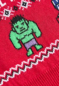Roter Strickstoff mit einer grünen Figur in lila Shorts, umrandet von schwarzer Linie, umgeben von dekorativen Mustern in Weiß und Schwarz.