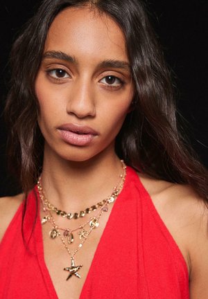 Jeune femme aux longs cheveux foncés portant plusieurs colliers en or et un haut rouge sans manches sur un fond noir.