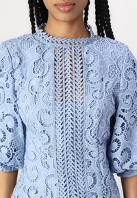 Lys blå blonde topp med intrikate blomster- og spiralmønstre, høy hals og knappelukking foran med vevd detalj.