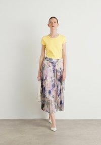 T-shirt en coton jaune à manches courtes, associé à une jupe maxi plissée à fleurs pastel. Le modèle porte des talons blancs et tient un petit sac.