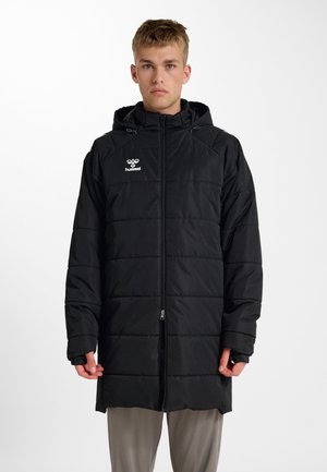 Ung mand stående iført en sort polstret lang jakke med hætte og et hvidt Hummel-logo på brystet, beige bukser.