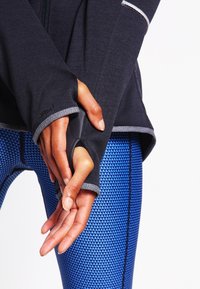 Zwarte zip-up top met duimgaten, gecombineerd met blauwe leggings met geometrisch patroon. Kenmerkt contraststiksel en textuurstoffen.