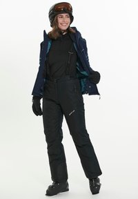 Mustat hiihtohousut säädettävillä olkaimilla, tummansinisestä kankaasta valmistettu takki turkoosilla vuorilla, yhdistettynä mustiin hansikkaisiin ja hiihtokengiin, sekä kypärä, jossa on suojalasit.