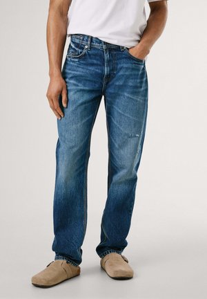 Vaqueros de mezclilla azul con un corte recto, que presentan detalles desgastados, costuras visibles y bolsillos frontales. Combinados con zapatos tipo slip-on en color beige.