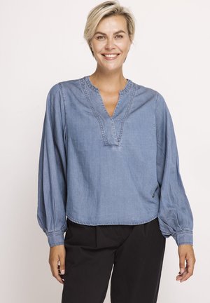 Lächelnde Frau mit kurzen blonden Haaren, die eine lockere hellblaue Langarmbluse und schwarze Hose vor einem einfarbigen Hintergrund trägt.