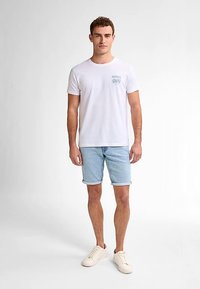 Camiseta blanca de algodón con un gráfico azul en el pecho, combinada con shorts de mezclilla azul claro y zapatillas beige. Look simple y casual.