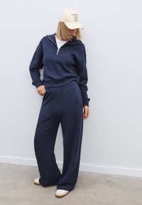 Námorníckomodrý sweatshirt s polovičným zipsom na golieri, ktorý je skombinovaný s širokými nohavicami v rovnakom odtieni. Hladká látka s elastickým pásom. Biele tenisky a svetlá čiapka.