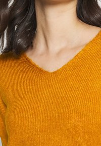 Stickad tröja i klar gul-orange med V-ringning. Har ribbad textur och en kroppsnära design. Inga synliga mönster eller hårdvara.