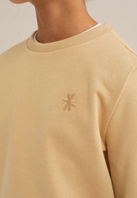 Minkštas, šviesiai geltonas crewneck tipo džemperis. Kairėje krūtinės pusėje yra mažas smėlio spalvos siuvinėtas gėlių motyvas. Lygus paviršius.