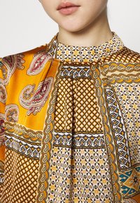 Blouse colorée avec des motifs cachemire et géométriques orange et crème. Tissu satin lisse avec un col montant et un design plissé.
