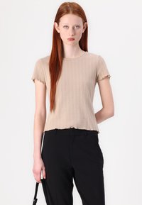 Beige t-skjorte med korte ermer, ribbestruktur og subtile vertikale mønstre, med rund hals og frynser, stylet med svarte bukser.