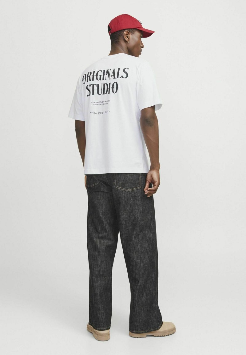 La camiseta blanca presenta un estampado negro de "ORIGINALS STUDIO" en la parte posterior. Combinada con unos pantalones de mezclilla oscuros y botas beige. Una gorra roja acentúa el look.