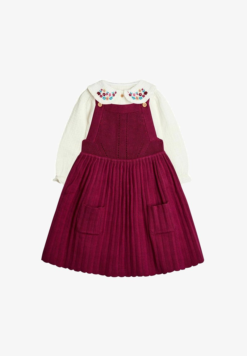 Camisa blanca de manga larga con cuello bordado de flores, combinada con un vestido de tirantes burdeos de punto que cuenta con bolsillos frontales y falda plisada.