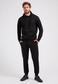 Schwarzes Hoodie- und Jogger-Set aus weichem Stoff. Das Hoodie hat eine Kängurutasche und eine Kordelzug; die Jogger haben elastische Bündchen und seitliche Taschen.