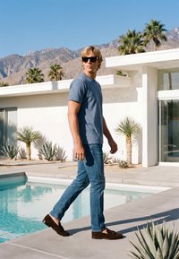 Homme blond avec des lunettes de soleil, chemise bleue et jean, marchant à côté d'une piscine près d'une maison blanche moderne avec des plantes désertiques et des montagnes en arrière-plan.