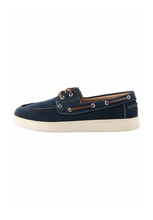 Chaussure bateau en daim bleu marine avec semelle blanche, lacets marron et coutures blanches, vue de côté sur fond blanc.