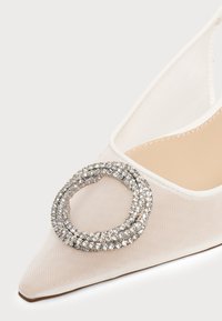 Gros plan d'une chaussure blanche à bout pointu avec un tissu en mesh et une grande boucle circulaire en strass sur le dessus.