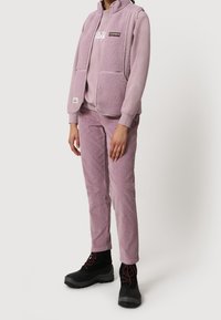 Chaleco de forro polar de color púrpura claro con bolsillos con cremallera, combinado con un sudadera de cuello redondo a juego y pantalones de pana. Botas de senderismo negras completan el conjunto.