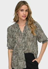 Camisa de botones de manga corta en tejido transparente con un estampado de leopardo en negro y beige. Presenta un cuello con volantes y un corte relajado.