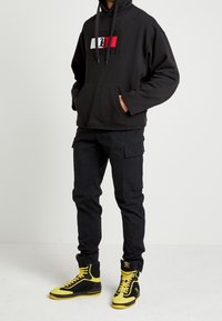 Zwarte hoodie met capuchon, voorzak en logo; zwarte cargobroek met zakken; zwart-gele hoge sneakers met veters.