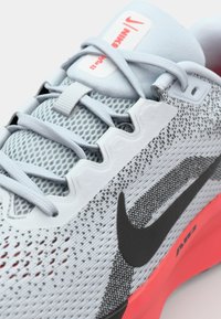 Chaussure de sport gris clair avec tige en mesh, logo Nike noir et semelle rouge vif. Motifs texturés notables et système de laçage discret.