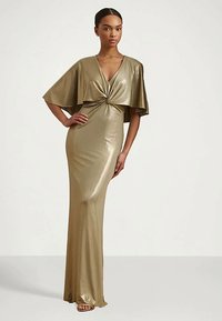 Γυναίκα που στέκεται φορώντας ένα μακρύ, γυαλιστερό χρυσό φόρεμα με στραμμένο μπροστά και φαρδιές κάπες κατά ενός απλού ανοιχτού φόντου.