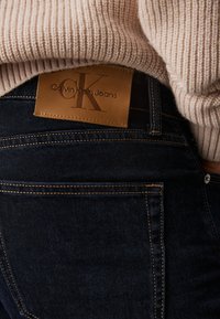 Jean en denim foncé avec une étiquette en cuir beige affichant le logo "Calvin Klein Jeans". Présente des coutures contrastées et un design ajusté.