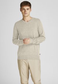 Suéter de cuello en V de manga larga color beige claro, hecho de material suave, con puños y dobladillo acanalados. Modelo combina con pantalones de color claro.
