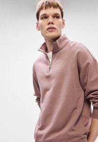 Bronzová mikina s vysokým límcem a zipovým uzávěrem, vybavená žebrovanými manžetami a diskrétním logem. Měkká textura a volný střih.