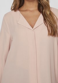 Blouse rose blush avec un col en V, des manches longues, et des détails de coutures minimalistes, présentant une coupe droite et une texture de tissu douce et lisse.