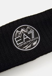 Tissu en tricot noir avec un patch rond noir et blanc du logo Emporio Armani EA7 présentant des montagnes et du texte.