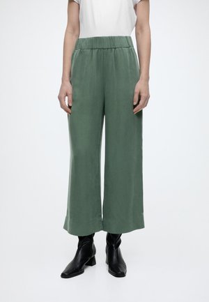 Personne portant un pantalon ample à jambes larges, taille haute, couleur vert olive, associé à des bottines noires à talons, sur fond blanc.