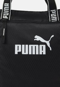 Czarna torba tote z nylonu, z fakturowaną powierzchnią, białym logo Puma oraz kontrastującymi paskami z tekstem "PUMA". Szczegół z zamkiem błyskawicznym.