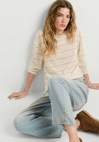 Femme aux longs cheveux ondulés assise par terre, portant une chemise beige rayée à manches longues, un jean bleu clair et des chaussures marron.